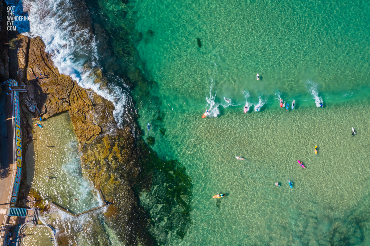 Aerial oceanscape above North Bondi Beach, with multi coloured surfboards catching a wave