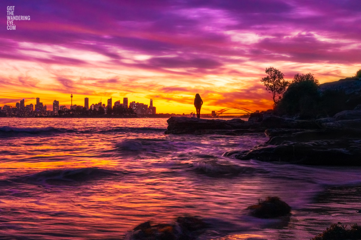 Woman standing admiring a stunning pink sky sunset from Milk Beach looking at the Sydney city skyline