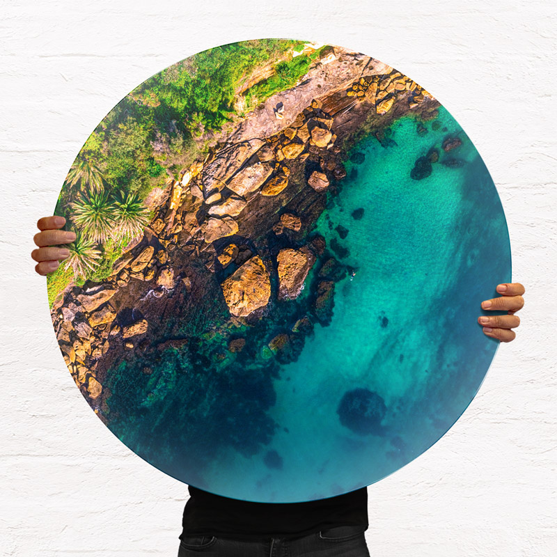 ChromaLuxe round metal prints of Sydneys beaches. Aerial oceanscape above the tranquil calm waters of Gordons Bay. Tropical palm trees as a swimmer enjoys a swim. Taken from the O'cean series of aerial prints by Allan Chan, Gotthewanderingeye. Wall art delivered and ready to hang.
