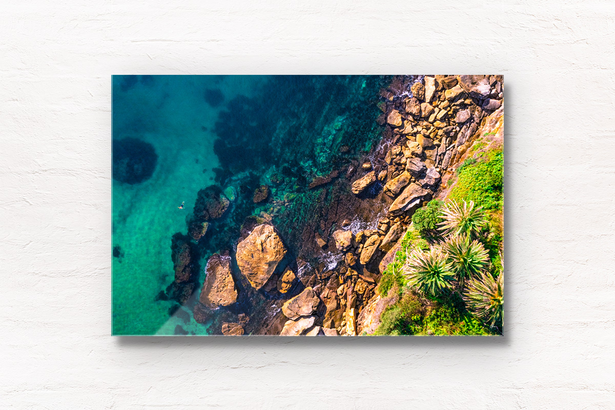 Framed wall art acrylic of Man Swimming in clear emerald waters of Gordons Bay, surrounded by palm trees and boulders from the clifftop.
