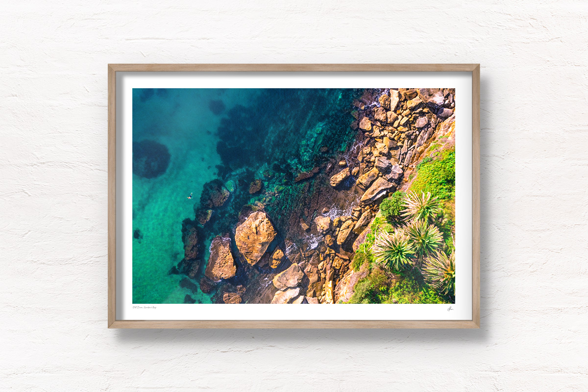 Framed wall art of man swimming in clear emerald waters of Gordons Bay, surrounded by palm trees and boulders from the clifftop.