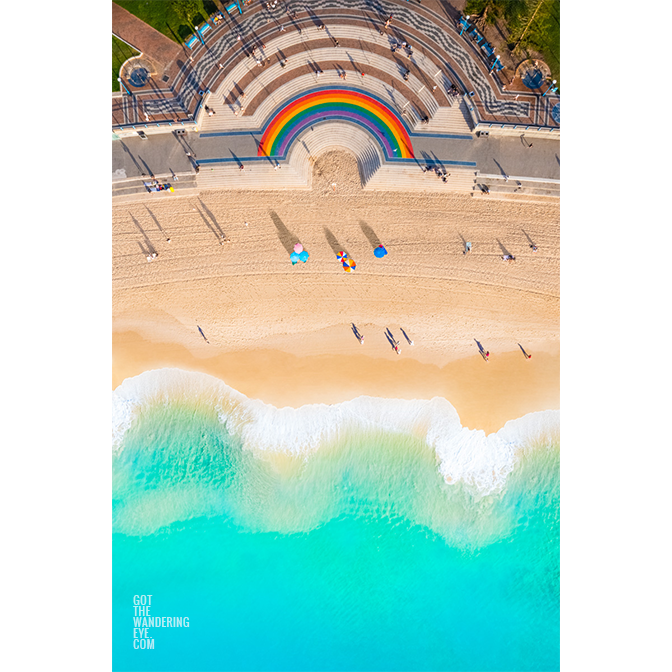 Vibrant 50 metre long rainbow on the promenade curving around the Coogee steps, as swimmers walk along the beach.