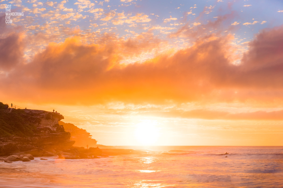 Golden warm light beaming through clouds on a Tamarama Beach Sydney Sunrise, lighting up Mackenzies point clifftops by Allan Chan.