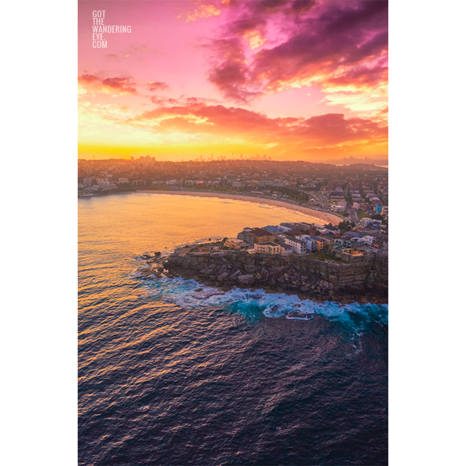 Aerial view above a Bondi Beach Sunset over the Sydney CBD skyline, glowing colour on the ocean waves.