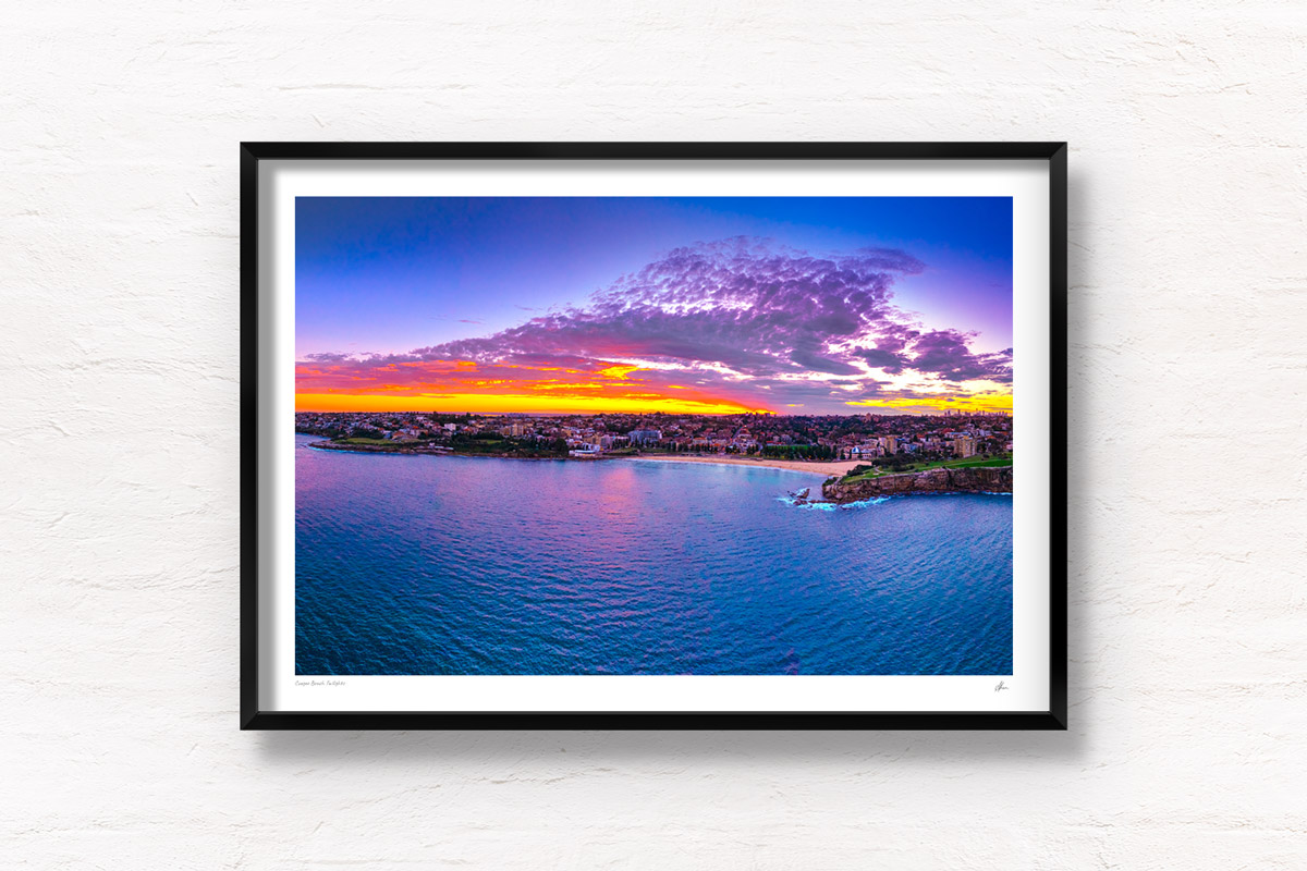 Aerial view above the ocean looking back at a Coogee Beach Sunset. Golden light through clouds with the Sydney CBD skyline on the horizon. Framed art photography wall art print.