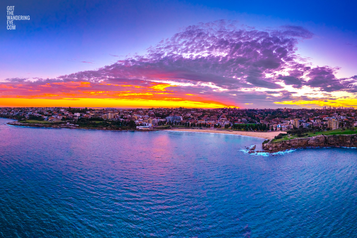 Aerial view above the ocean looking back at a Coogee Beach Sunset. Golden light through clouds with the Sydney CBD skyline on the horizon.