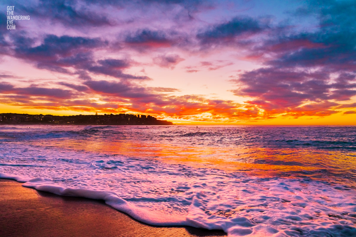 Close up of frothy waves rolling onto Bondi Beach as a fiery red colourful sunrise appears over Ben Buckler.