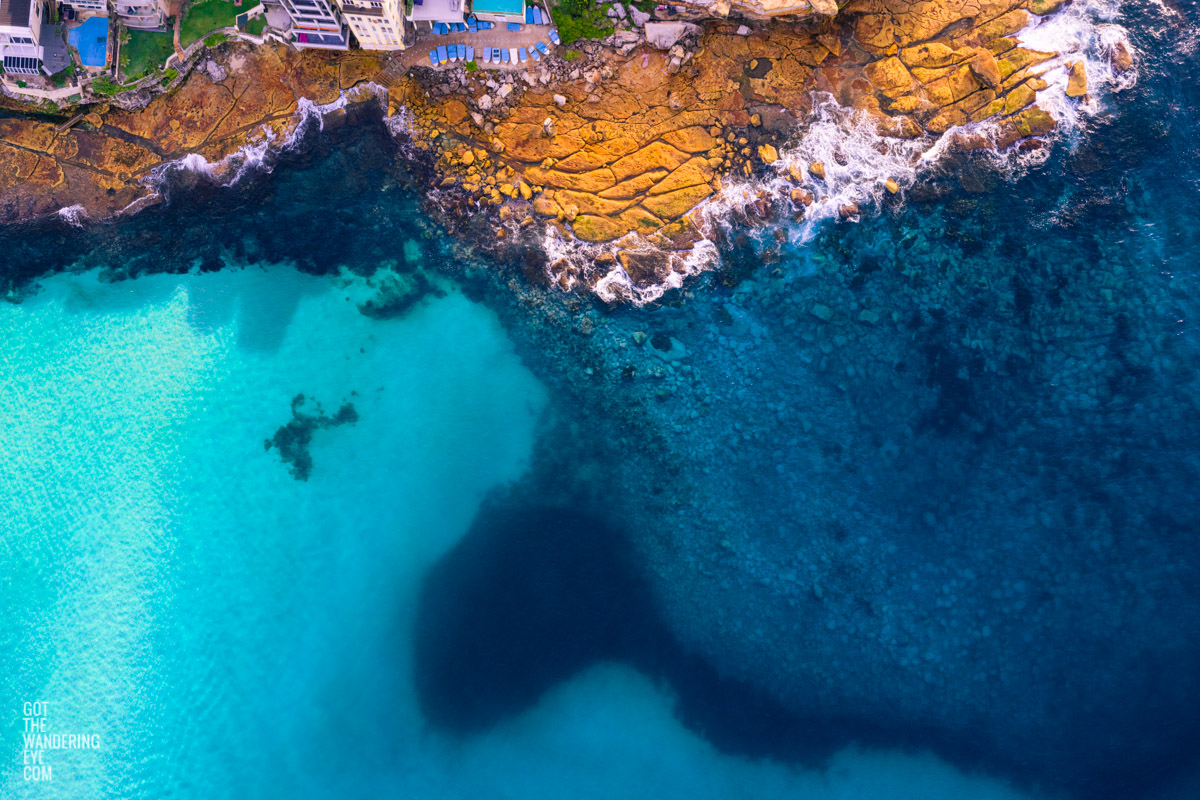 Aerial view above the blues of north Bondi Boat sheds and crystal clear ocean surrounding flat rock.