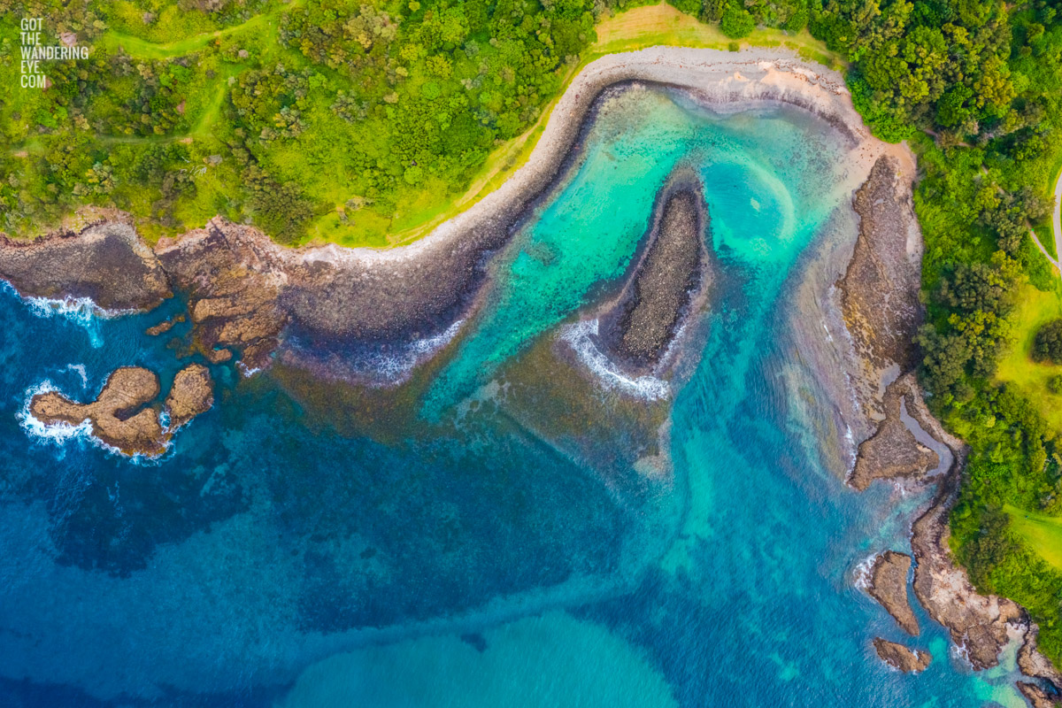 Aerial view above a secluded pebbly boneyard beach, Kiama Downs. Teal coloured water surrounded by trees.