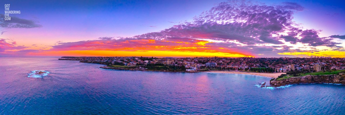 Aerial view above the ocean looking back at a Coogee Beach Sunset. Golden light through clouds with the Sydney CBD skyline on the horizon.