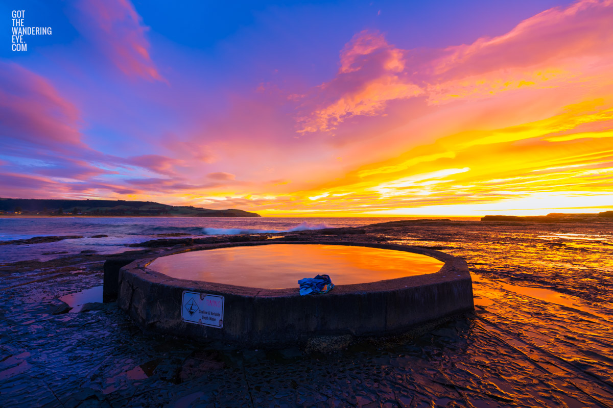 Purple and orange sunrise fluffy clouds above Werri Beach and round ocean rock pool.